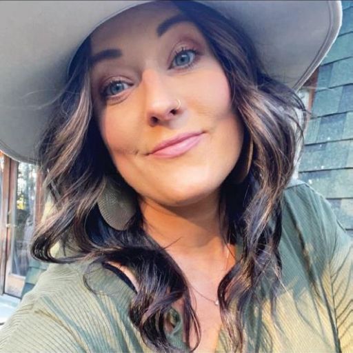 A woman with long wavy brown hair, wearing a wide-brimmed hat, green top, and hoop earrings, smiles at the camera in an outdoor setting with sunlight and greenery—radiating the spirit of Restoring Hope like Malerie Lockhart.