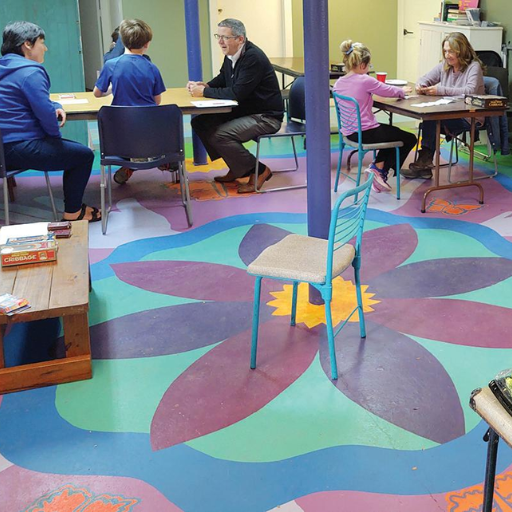 Adults and children sit at tables playing games in a colorful Recovery Community Center, with a large painted flower design on the floor and board game boxes nearby. The atmosphere is relaxed and focused.