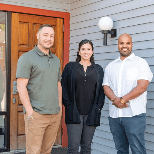 Three people stand smiling in front of a gray building with wood door and glass window, possibly outside a SaVida Health center. Two men, one in green and one in white, stand on either side of a woman in black—highlighting the support of addiction treatment.