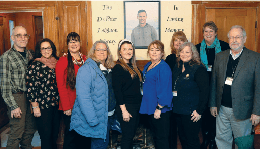 A group of ten adults stands indoors, smiling at the camera in front of a memorial sign that reads, The Dr. Peter Leighton Library In Loving Memory, at Tuckers House in Bridgton, with a framed photo of a man displayed above them.