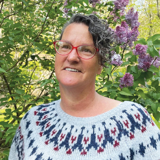 A person with short curly gray hair and red glasses stands outdoors in front of blooming lilac bushes, wearing a light-colored sweater with a dark blue and red pattern—OPTIONS Liaison Dawn Kearns enjoys the spring day.