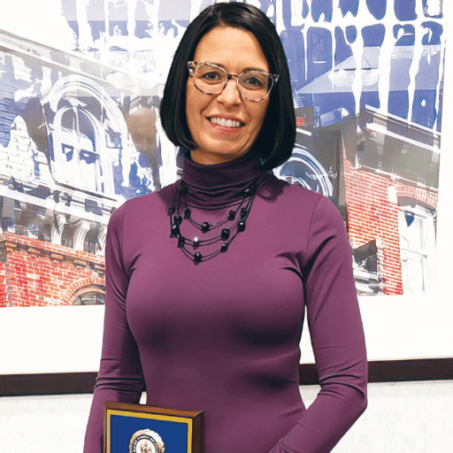 A woman with shoulder-length dark hair and glasses, wearing a purple turtleneck and a black beaded necklace, stands indoors holding a plaque for the Liaison Spotlight. A large, partially blurred building photograph is on the wall behind her.