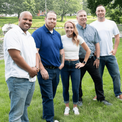 Five adults stand together on green grass by a river, smiling at the camera. Casually dressed in jeans and t-shirts, they reflect the positive spirit of recovery. Trees and a small white building appear in the background.