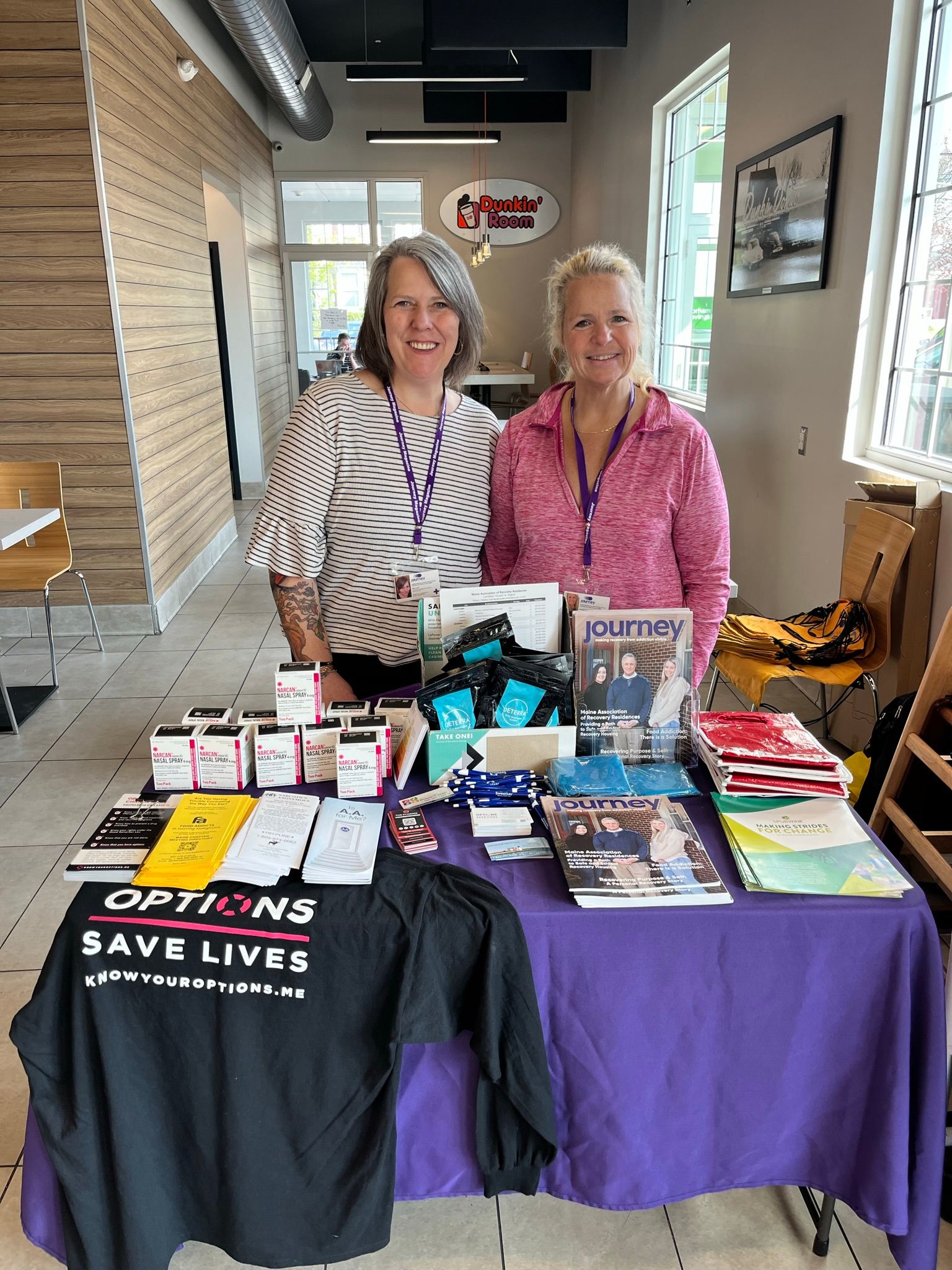 Two women from Maine Treatment stand behind an informational table with brochures, magazines, naloxone kits, and a shirt reading “OPTIONS SAVE LIVES” in a bright room. Both are smiling and wearing purple lanyards, supporting recovery in their community.