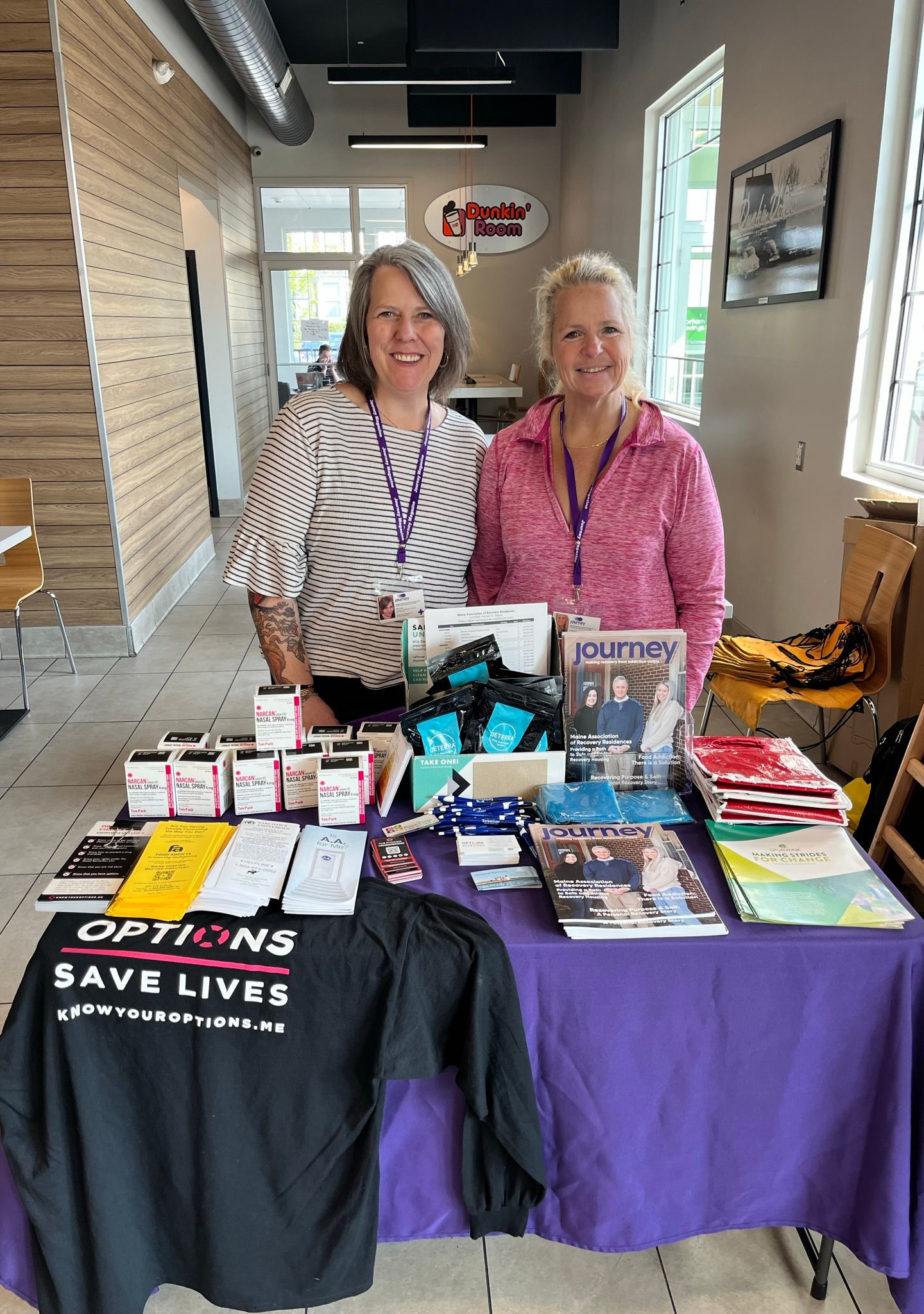 Two women from Maine Treatment stand behind an informational table with brochures, magazines, naloxone kits, and a shirt reading “OPTIONS SAVE LIVES” in a bright room. Both are smiling and wearing purple lanyards, supporting recovery in their community.