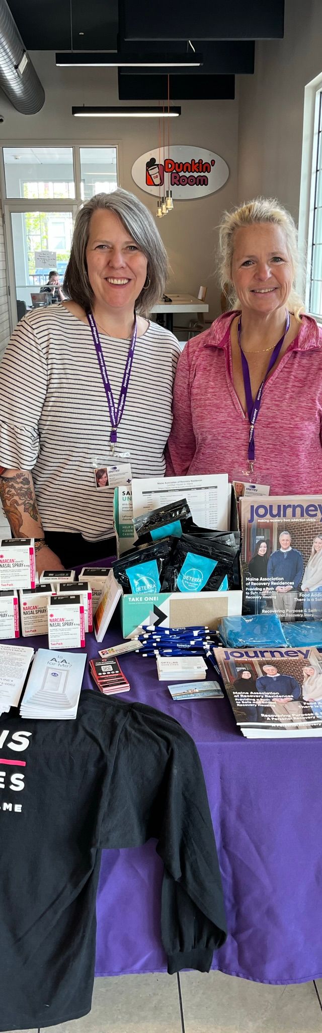 Two women from Maine Treatment stand behind an informational table with brochures, magazines, naloxone kits, and a shirt reading “OPTIONS SAVE LIVES” in a bright room. Both are smiling and wearing purple lanyards, supporting recovery in their community.