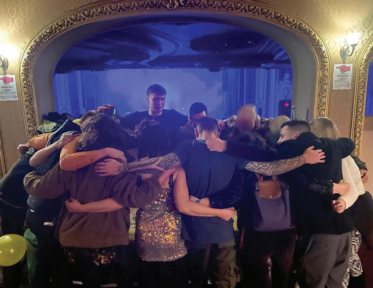 A group of people stand in a circle with their arms around each other, facing inward under ornate lighting, sharing a moment of togetherness in a warmly-lit room with a stage and blue-lit backdrop—a true sober social scene.