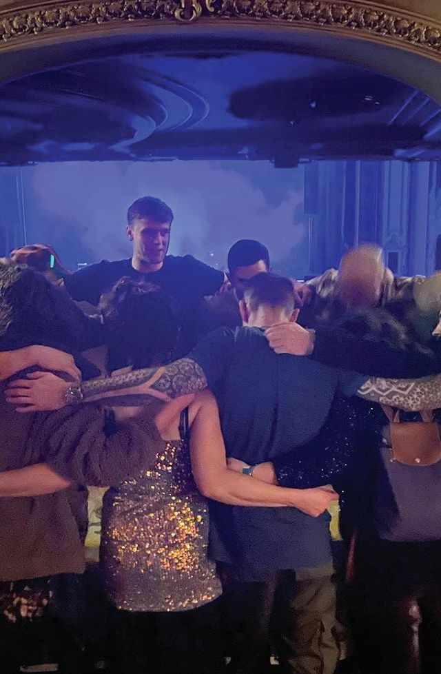 A group of people stand in a circle with their arms around each other, facing inward under ornate lighting, sharing a moment of togetherness in a warmly-lit room with a stage and blue-lit backdrop—a true sober social scene.
