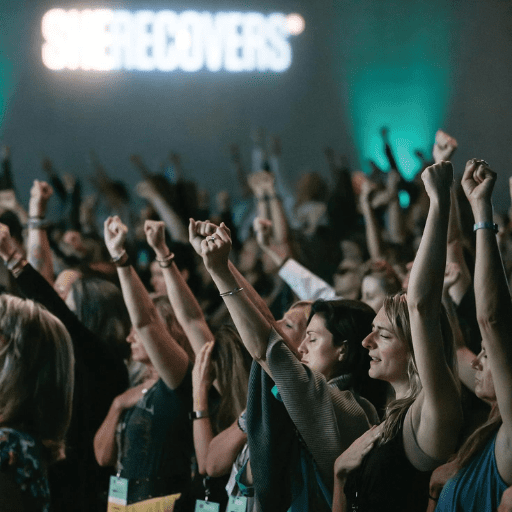 A large group of people at an indoor She Recovers event stand with their arms raised in solidarity. The background features a blurred sign that reads SHERECOVERS and teal-colored lighting.