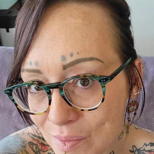 A person with glasses, facial piercings, and several tattoos is looking at the camera. Embodying a unique journey, Crystale Vega has drawn or tattooed dots above their eyebrows and is wearing earrings.