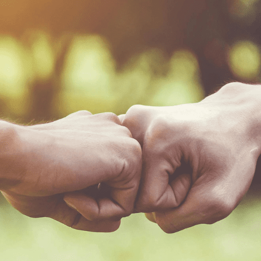 Two people, possibly Peer Recovery Coaches, bump fists in a friendly gesture, with a blurred outdoor background and warm lighting.