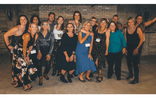 A group of people, mostly women, stand closely together indoors, smiling and posing for a photo against a brick wall. Dressed in semi-formal attire, they appear to be attending a Sober Social Society celebratory event.