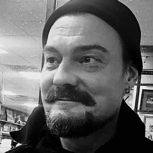 Black and white photo of a man with a mustache and goatee, wearing a beanie and earrings, smiling slightly. His inked and sober journey shows in his eyes, hinting at stories of restoring hope. Framed pictures and ceiling lights fill the background.