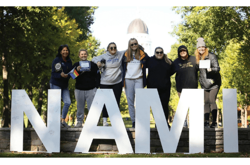 Seven people stand outdoors behind large white letters spelling NAMI, smiling and posing together at a NAMIWalks Maine event. Trees line the background, and a domed building is visible in the distance—promoting mental health awareness in Maine.