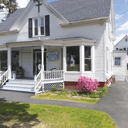 Friendship House is a white, two-story home with a front porch, black shutters, and a cross above the door. A blooming pink shrub sits by the foundation, and a paved driveway curves in front on a sunny day.