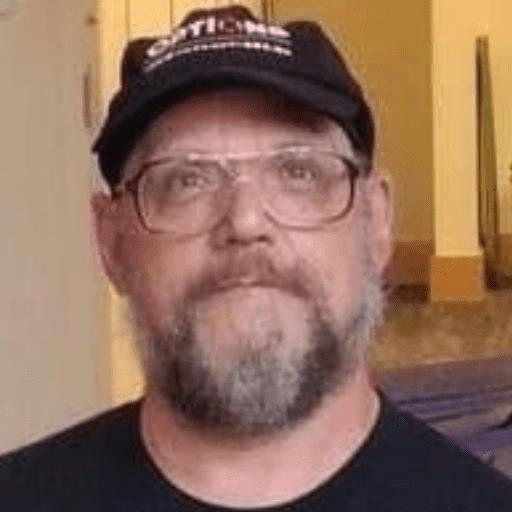 Dave Bilodeau, an OPTIONS Liaison, is a bearded man with glasses and a black cap who looks slightly upward. He wears a black shirt against a softly lit background of yellow and beige tones.
