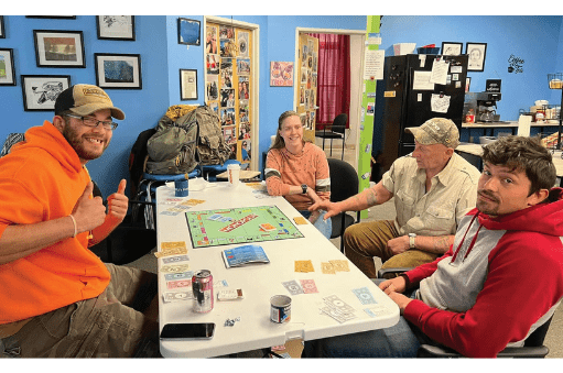 Four adults sit around a white table at Inspire Recovery Center in Ellsworth, playing Monopoly in a colorful room. One man in an orange hoodie gives a thumbs up and smiles while the others focus on the game, with drinks and game money on the table.