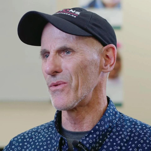 A middle-aged man with light skin, wearing a black baseball cap and a blue patterned shirt, looks slightly to the side while speaking. The background is blurred, suggesting a connection to Maine’s OPTIONS Liaisons program.