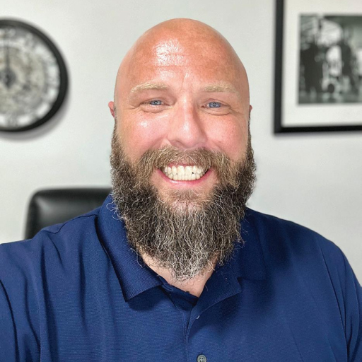 Josh Colwell, a smiling bald man with a full beard, sits indoors in a navy blue collared shirt, with a black-and-white photo and a clock on the wall behind him.