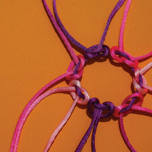 Close-up of pink and purple plastic lacing cords tied in knots on an orange background, their vibrant fibers woven together like threads of community, evoking friendship bracelets or classic lanyard crafts.