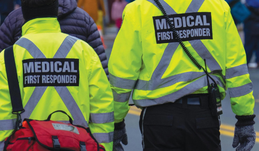 Two medical first responders in bright yellow jackets with reflective stripes, part of a Maine EMS SUD Response team, walk side by side—one carrying a red medical bag—on a street with other people visible in the blurred background.