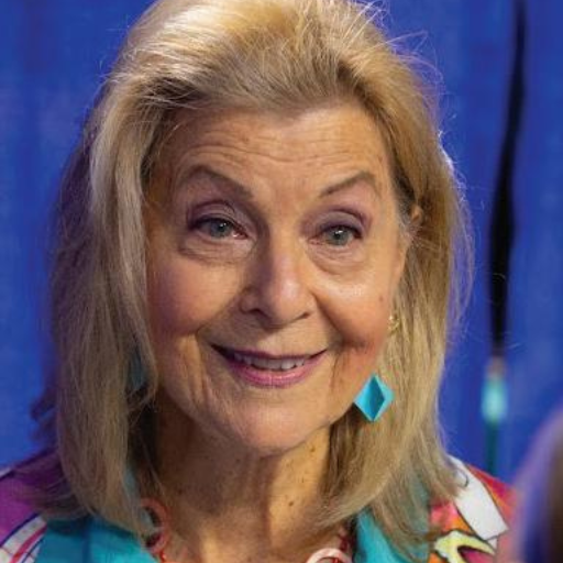 An older woman with blonde hair smiles warmly, embodying the spirit of discovery. She wears turquoise earrings and a colorful top, with a blue curtain in the background.