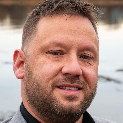 A man with short brown hair and a trimmed beard, wearing a gray suit jacket, smiles slightly while standing outdoors near a body of water.