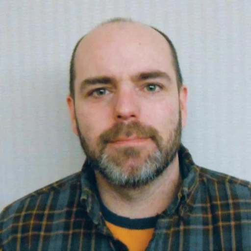 A man with a beard and mustache wearing a plaid shirt looks at the camera with a neutral expression. The plain, light background puts the spotlight on this Somerset County New Options Liaison.