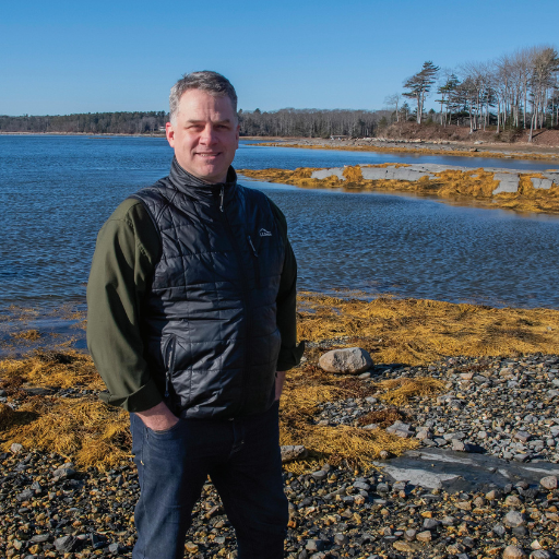 A man stands on a rocky, seaweed-covered shore with his hands in his pockets, wearing a dark vest and green sweater. Under a clear blue sky, Matt Senecal reflects by the calm water and trees—capturing a quiet moment of recovery.