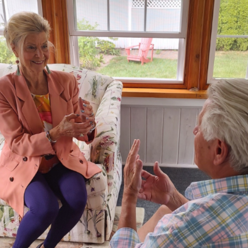 An older woman and man sit facing each other indoors, smiling and gesturing with their hands as if playing a game. The woman sits on a floral couch; the man sits on a chair. Sunlight streams in through large windows behind them.