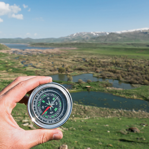 Person holds a compass in their hand, with a valley, river, and snow-capped mountains visible in the background.