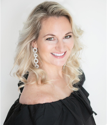Woman with blonde wavy hair wearing black off-shoulder top and dangling silver chain earrings, smiling at camera.