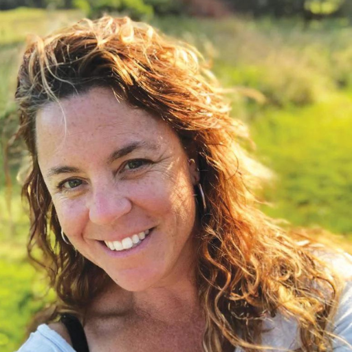 Woman with reddish-brown curly hair smiling outdoors with green grass background.