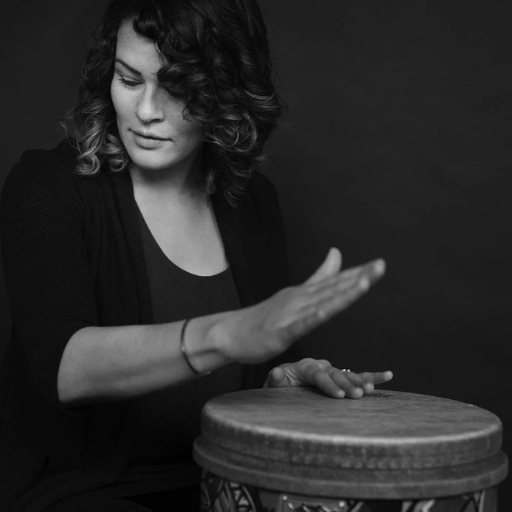 Person with wavy shoulder-length hair plays a hand drum against a dark background.