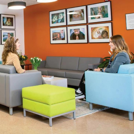 Two people sit in comfortable chairs in a bright orange room with framed photos on the wall, facing each other across a lime green ottoman.