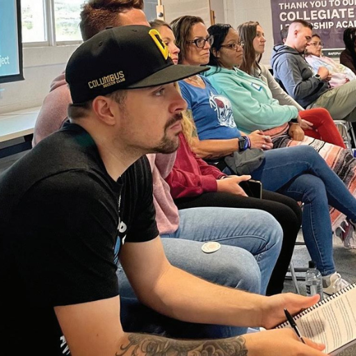 Six people sit in a row at an indoor event, listening attentively to a speaker.