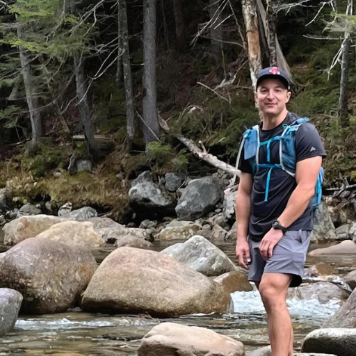 Man wearing blue backpack and cap stands on rocks beside a forest stream.