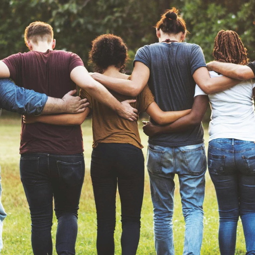 Four people stand in a circle facing away from the camera with arms around each other's shoulders in a grassy outdoor setting.