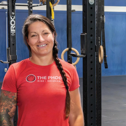 Woman with braided hair wearing red The Phoenix Rise Recovery shirt stands in front of gym equipment.