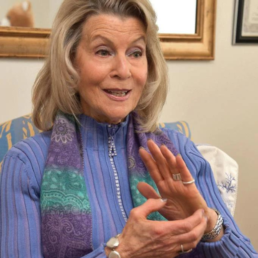 Woman in blue blouse gesturing with hands while speaking in an indoor room with framed artwork.