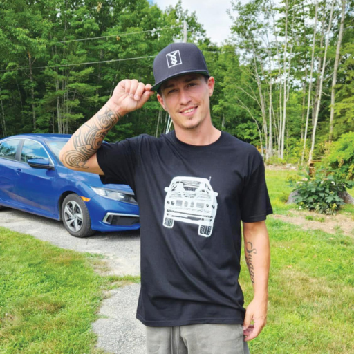 Man wearing black cap and car-graphic shirt stands in driveway with blue sedan and trees behind him.