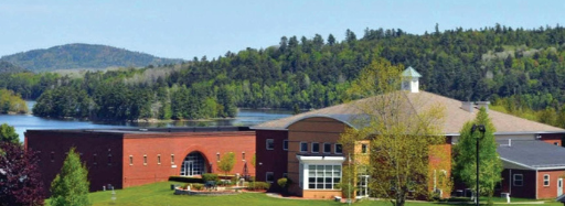 Modern brick buildings with large windows sit on a grassy campus surrounded by forested hills and a lake.