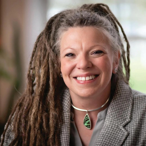 Woman with long dreadlocks wearing a gray jacket and pendant necklace smiles at camera.