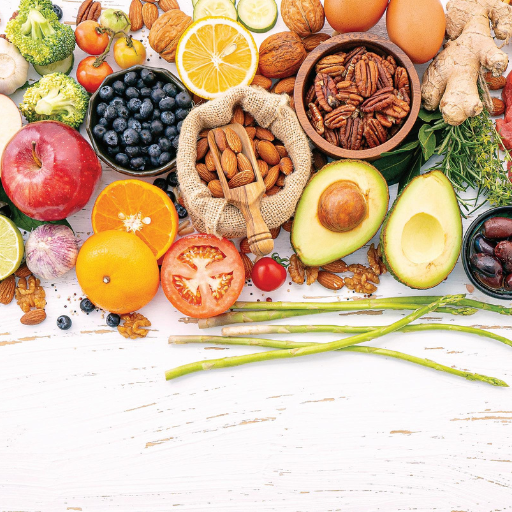 Colorful assortment of fresh vegetables, fruits, nuts, and grains arranged on white wooden surface.