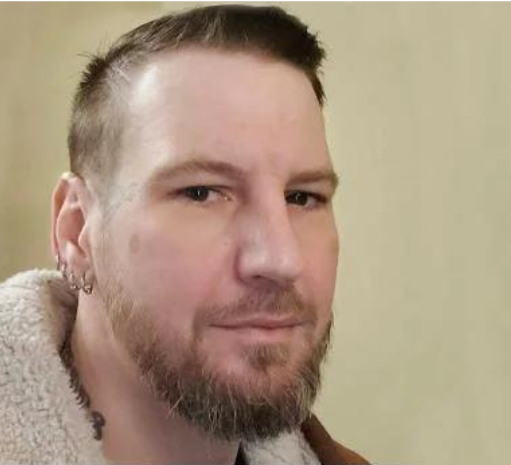 Man with beard and short dark hair wearing a tan sweater, facing camera against a beige wall.