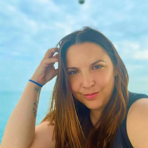 Woman with long brown hair touching her head, smiling at camera with ocean and blue sky behind her.