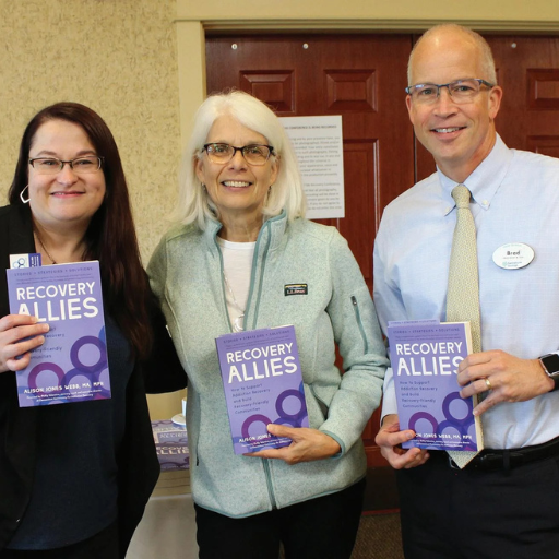 Three people stand indoors holding purple Recovery Allies books at what appears to be an event or presentation.