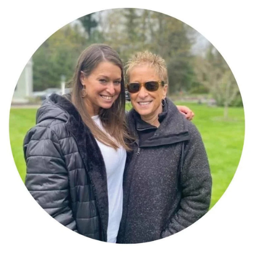 Two women smile at the camera while standing in a grassy park with trees behind them.