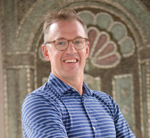 Man with glasses wearing blue striped polo shirt smiles in front of ornate stone archway.