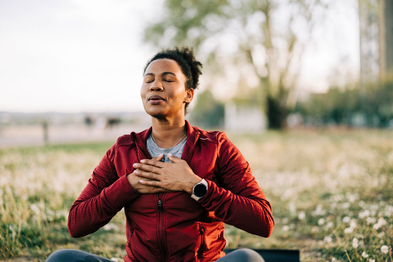 A person wearing a red jacket sits outdoors on grass with eyes closed, hands placed on their chest, appearing calm and relaxed—possibly meditating, practicing mindfulness, or focusing on mental health and wellness.